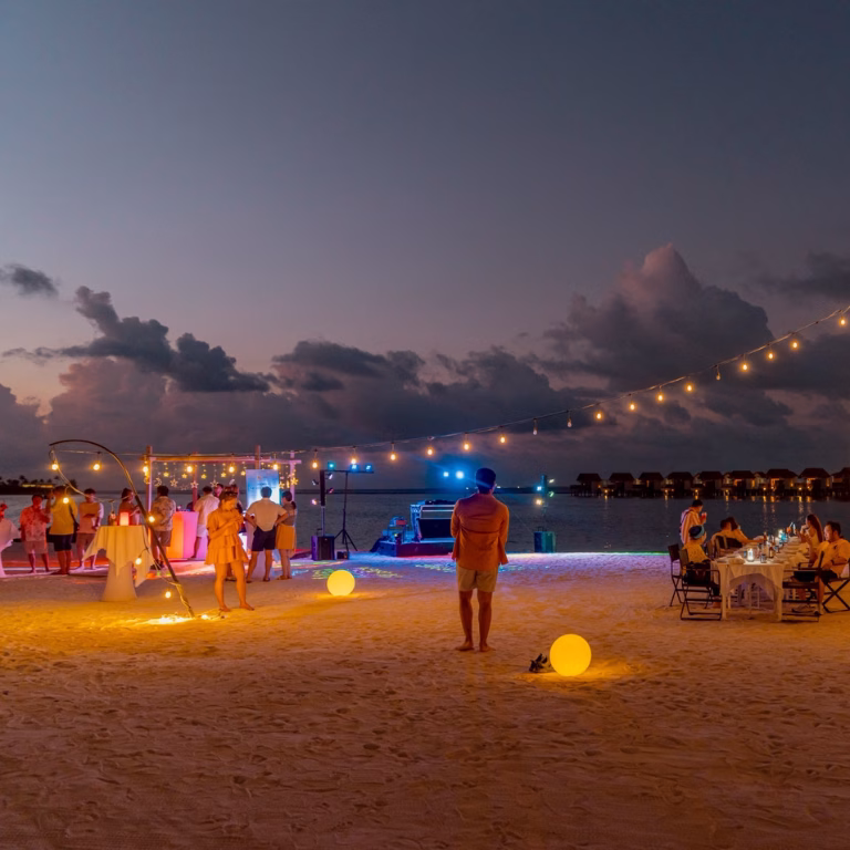 บรรยากาศงานเลี้ยงอาหารค่ำแบบส่วนตัวบนชายหาด (Private Beach Dinner) ยามค่ำคืน ตกแต่งด้วยไฟประดับ โต๊ะอาหารบนทราย และบอลไฟเรืองแสง สำหรับคณะ Gilenfah