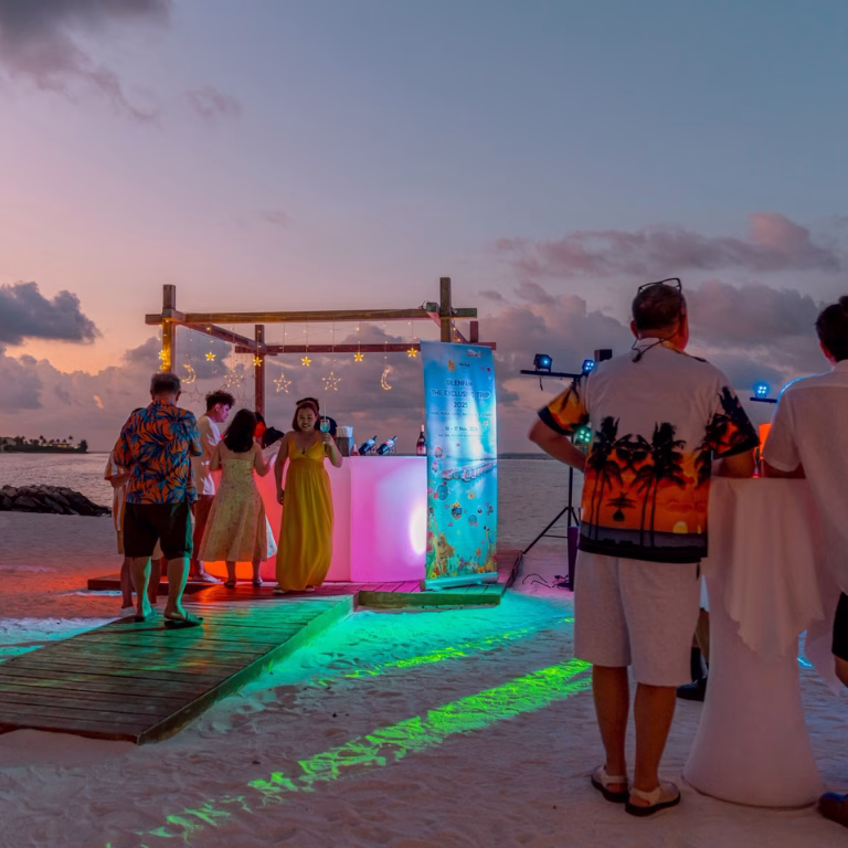 บรรยากาศงานเลี้ยงสังสรรค์ริมหาดช่วงพระอาทิตย์ตก (Sunset Party) พร้อมซุ้มบาร์เครื่องดื่มเรืองแสงและป้ายต้อนรับคณะ Gilenfah Exclusive Trip 2025