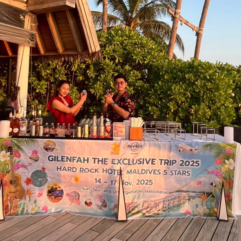 บาร์เทนเดอร์เตรียมเครื่องดื่มต้อนรับที่ซุ้มบาร์ริมหาด พร้อมป้ายไวนิล Gilenfah The Exclusive Trip 2025 ในบรรยากาศปาร์ตี้ช่วงพระอาทิตย์ตก